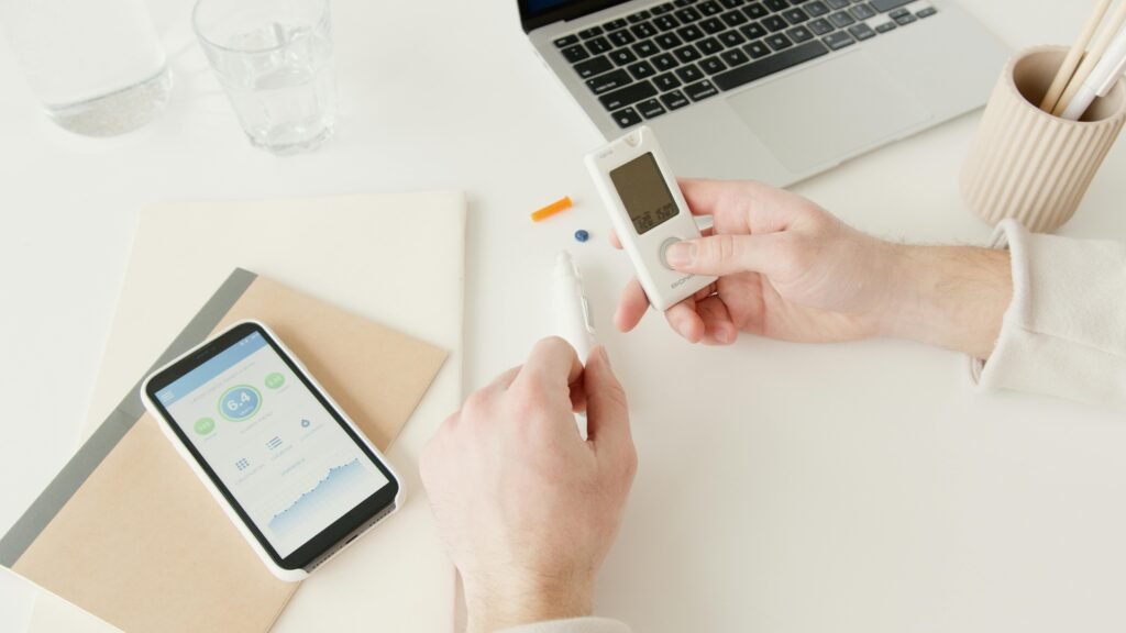 Person checking blood sugar with glucometer on a desk with smartphone and laptop nearby.